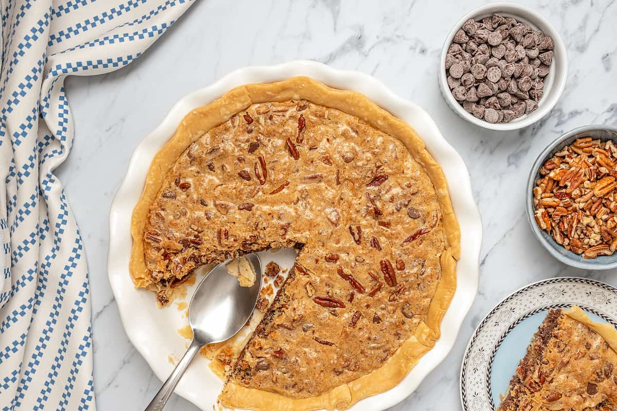 Gluten-free chocolate chip cookie pie with a slice missing, next to small bowls of chocolate chips and pecans.