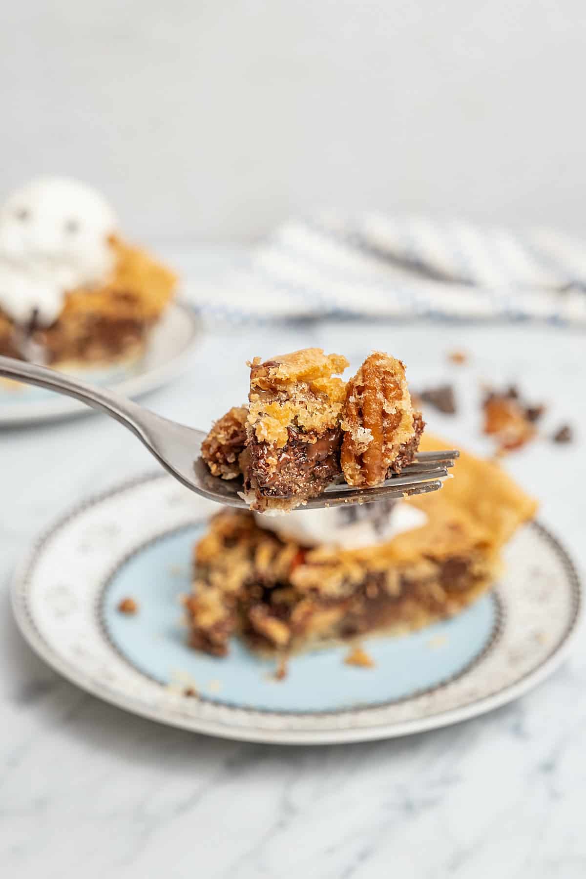 Close up of a forkful of chocolate chip cookie pie with a slice of pie on a plate in the background.