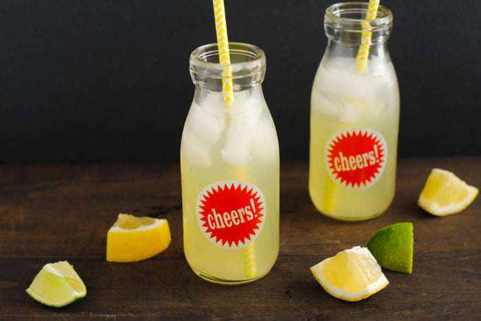 Glass bottles of agave lemonade with straws next to scattered lemon and lime wedges.