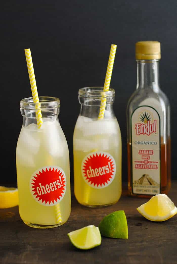 Glass bottles of agave lemonade with straws, next to a bottle of agave nectar and scattered lemon and lime wedges.