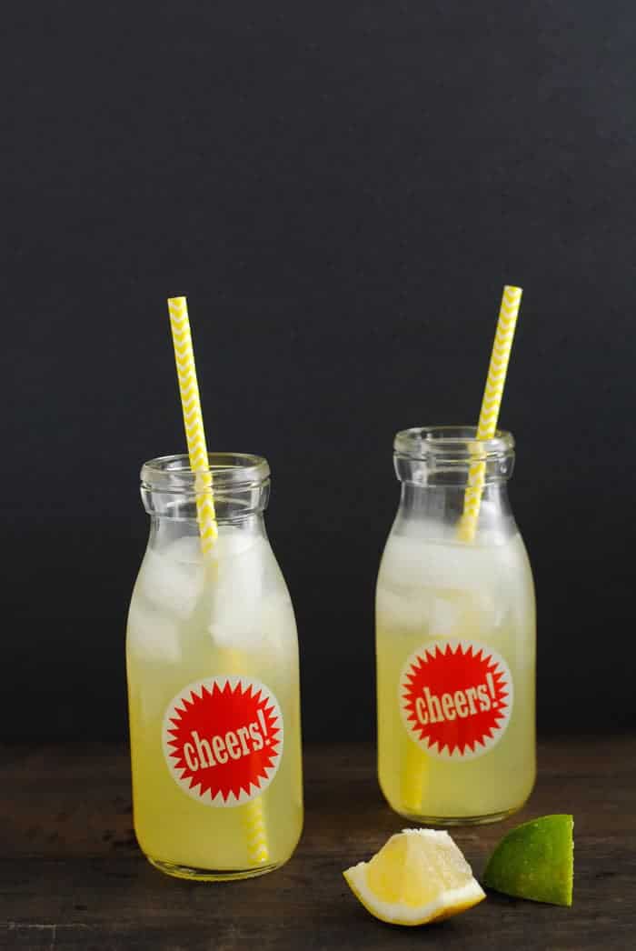 Glass bottles of agave lemonade with straws next to scattered lemon and lime wedges.