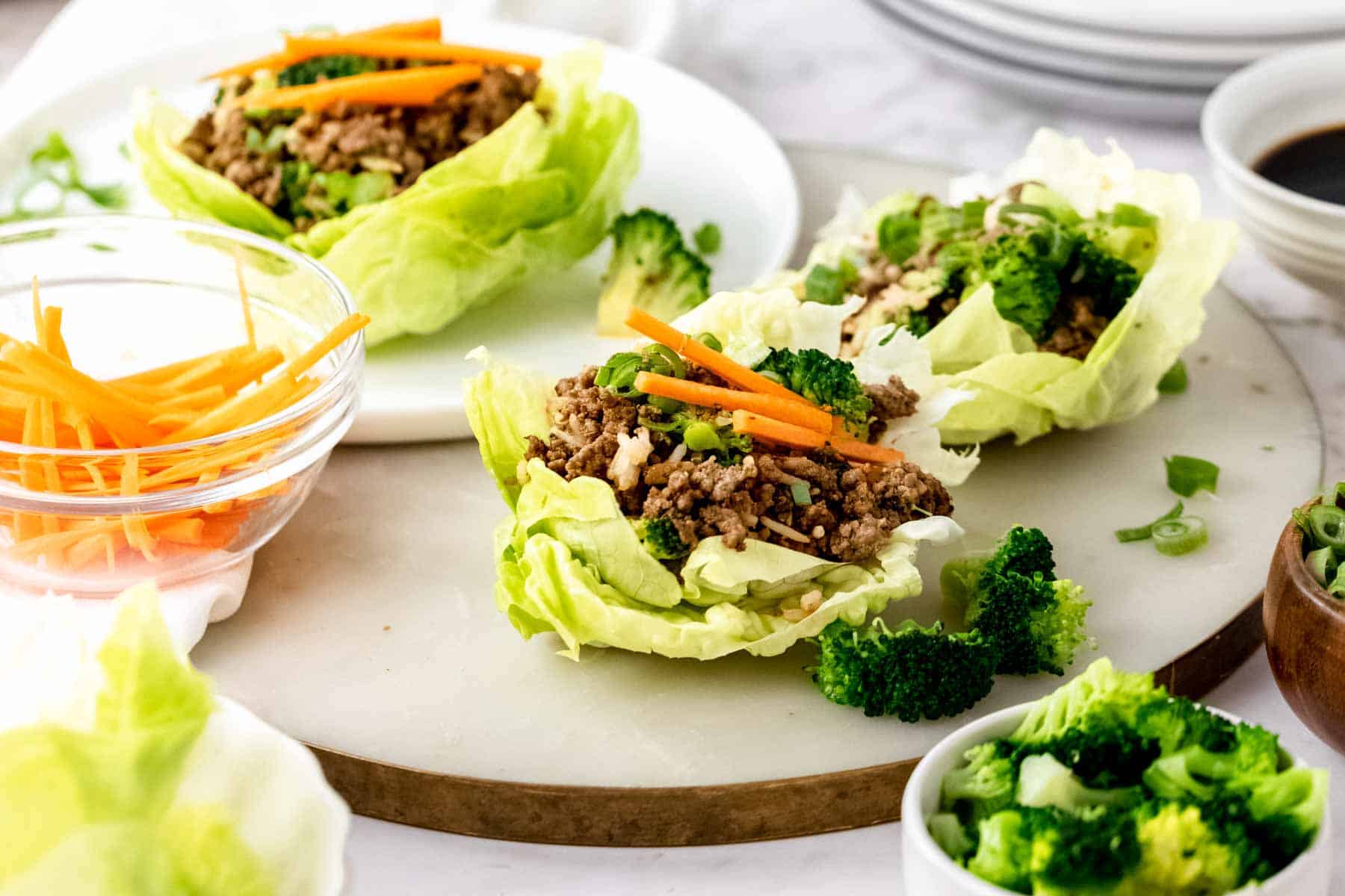 Two Asian beef lettuce wraps on a countertop next to a third lettuce wrap on a white plate, surrounded by wrap ingredients.