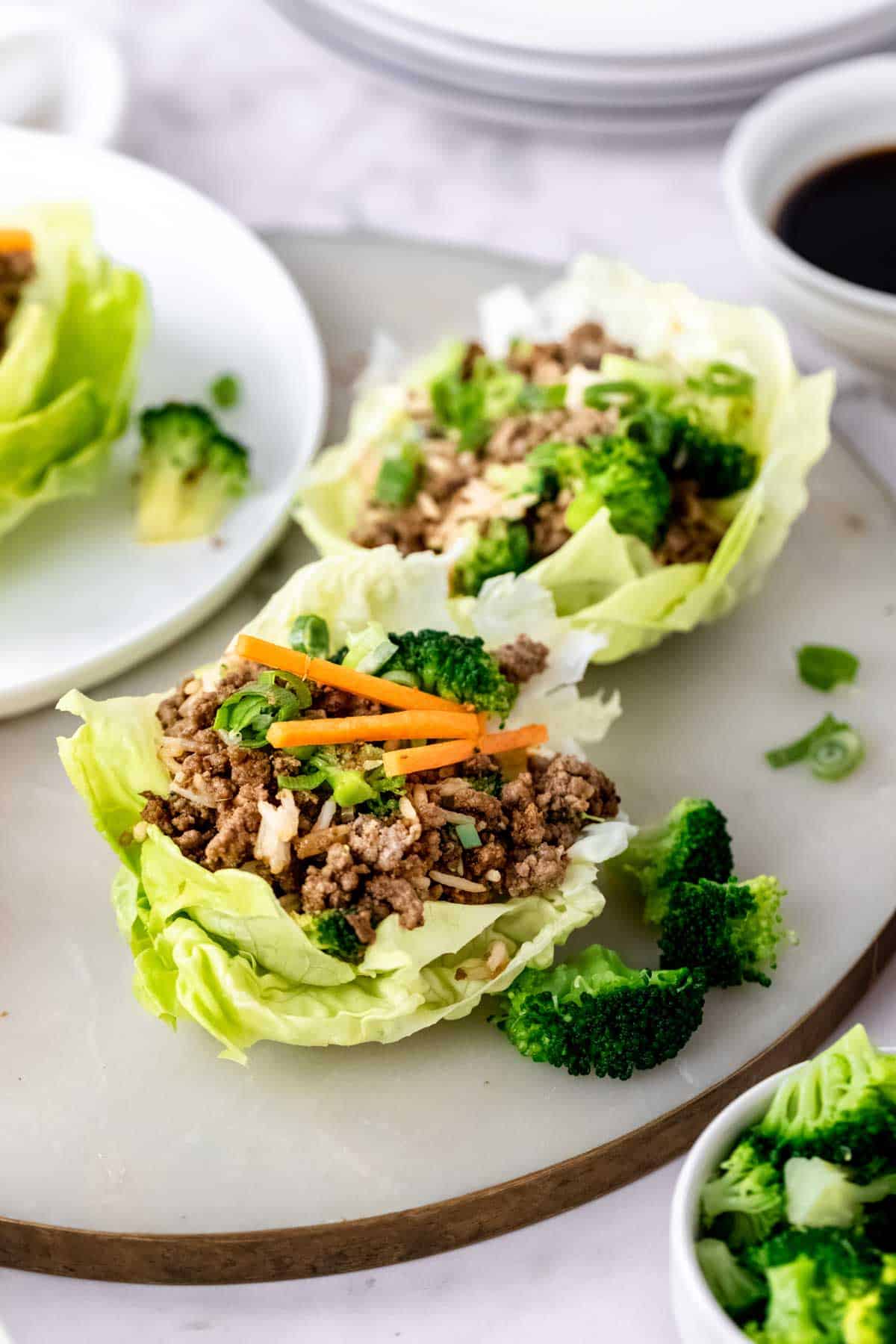 Two Asian beef lettuce wraps on a countertop next to a third lettuce wrap on a white plate.