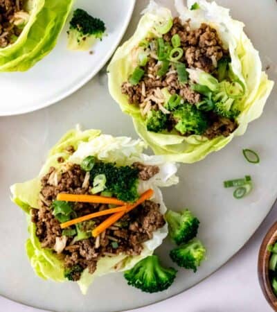 Overhead view of two Asian beef lettuce wraps on a countertop next to a third lettuce wrap on a white plate.