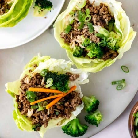 Overhead view of two Asian beef lettuce wraps on a countertop next to a third lettuce wrap on a white plate.