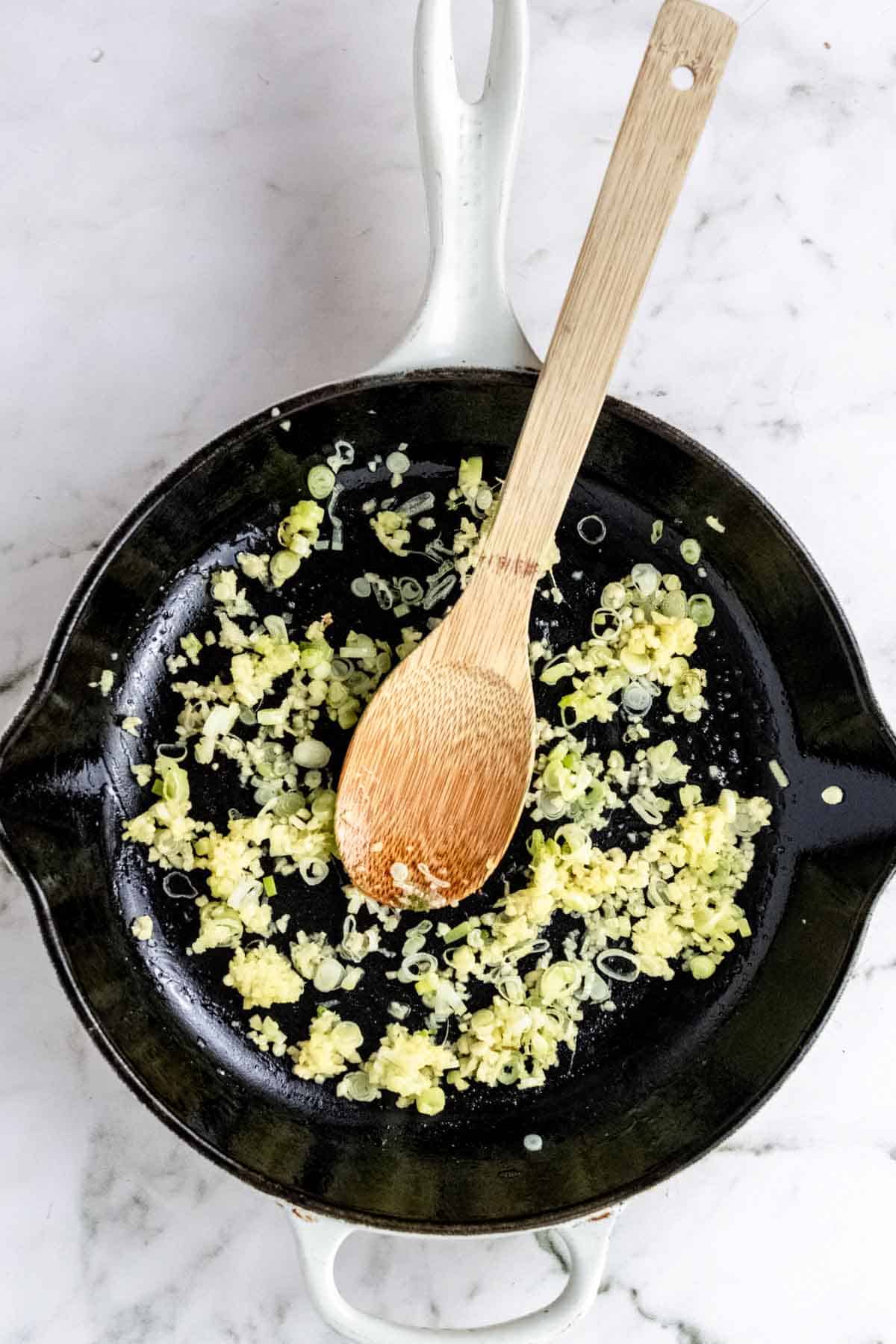 Scallions sauteing with garlic and ginger in a skillet with a wooden stirring spoon.