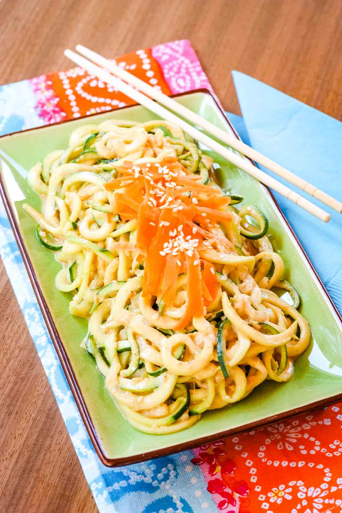 Cold Sesame Zoodles Salad on a light green rectangular plate on top of a colorful napkin with chopsticks resting on the side of the dish.