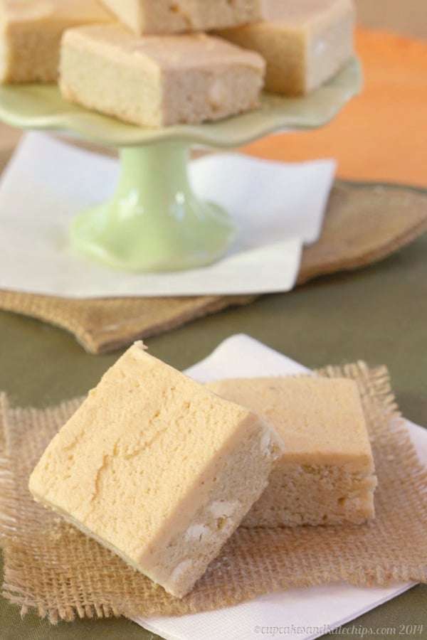 Two frosted sugar cookie bars in the foreground with a tray full of cookie bars in the background