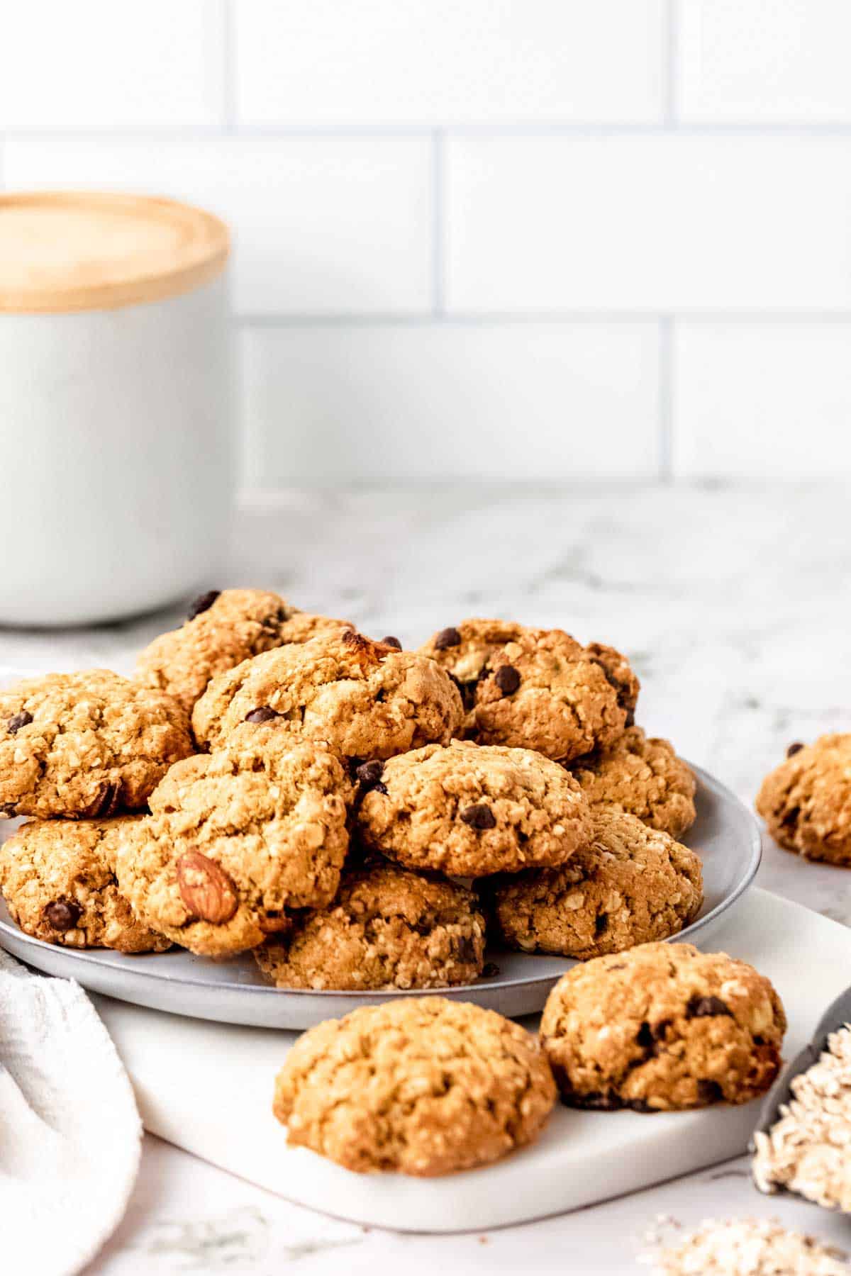 Assorted gluten-free trail mix cookies piled on a grey plate, on a white marble countertop with a cookie jar in the background.