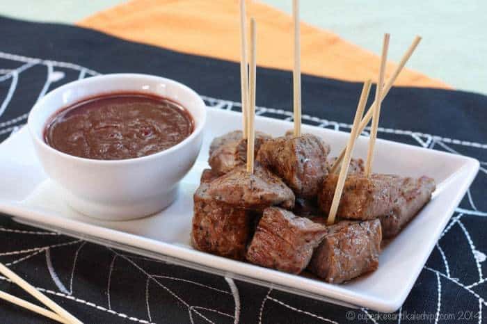 A white platter with steak bites on skewers and a red wine tomato dipping sauce