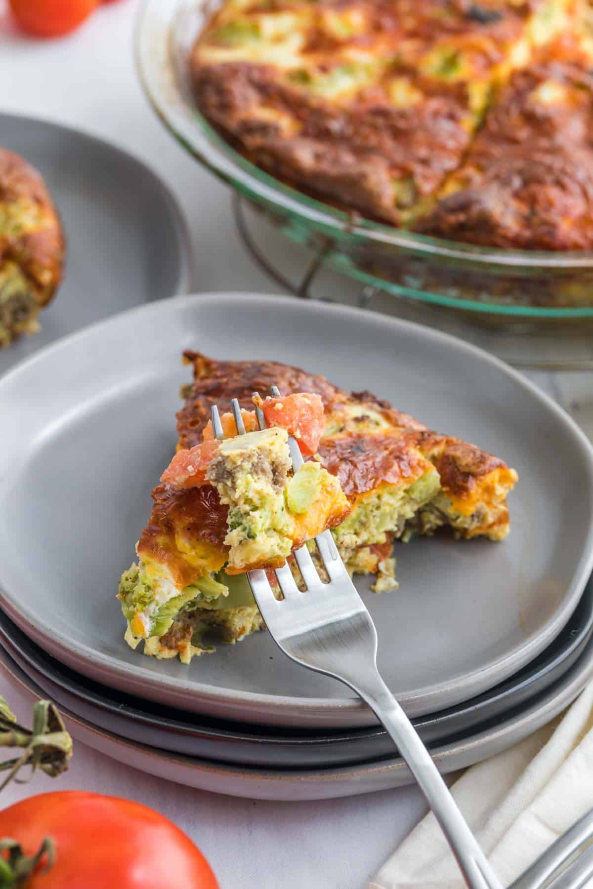 A slice of tomato broccoli sausage quiche on plate with a work that has a bite of the quiche on it.