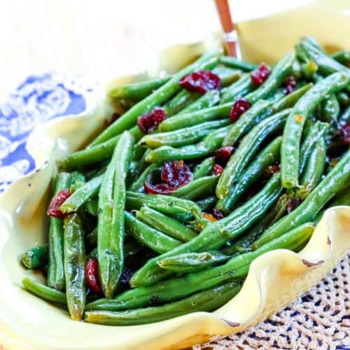 Orange glazed green beans with dried cranberries in a yellow serving dish with a spoon handle sticking out.