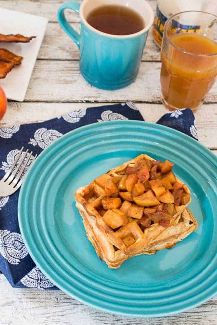 Apple cider waffles topped with caramelized apples on a blue plate, next to a mug of coffee.