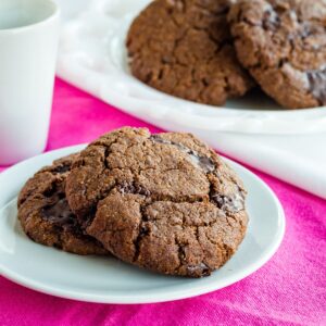 Two chocolate chunk cookies on a small white plate on top of a pink cloth napkin.