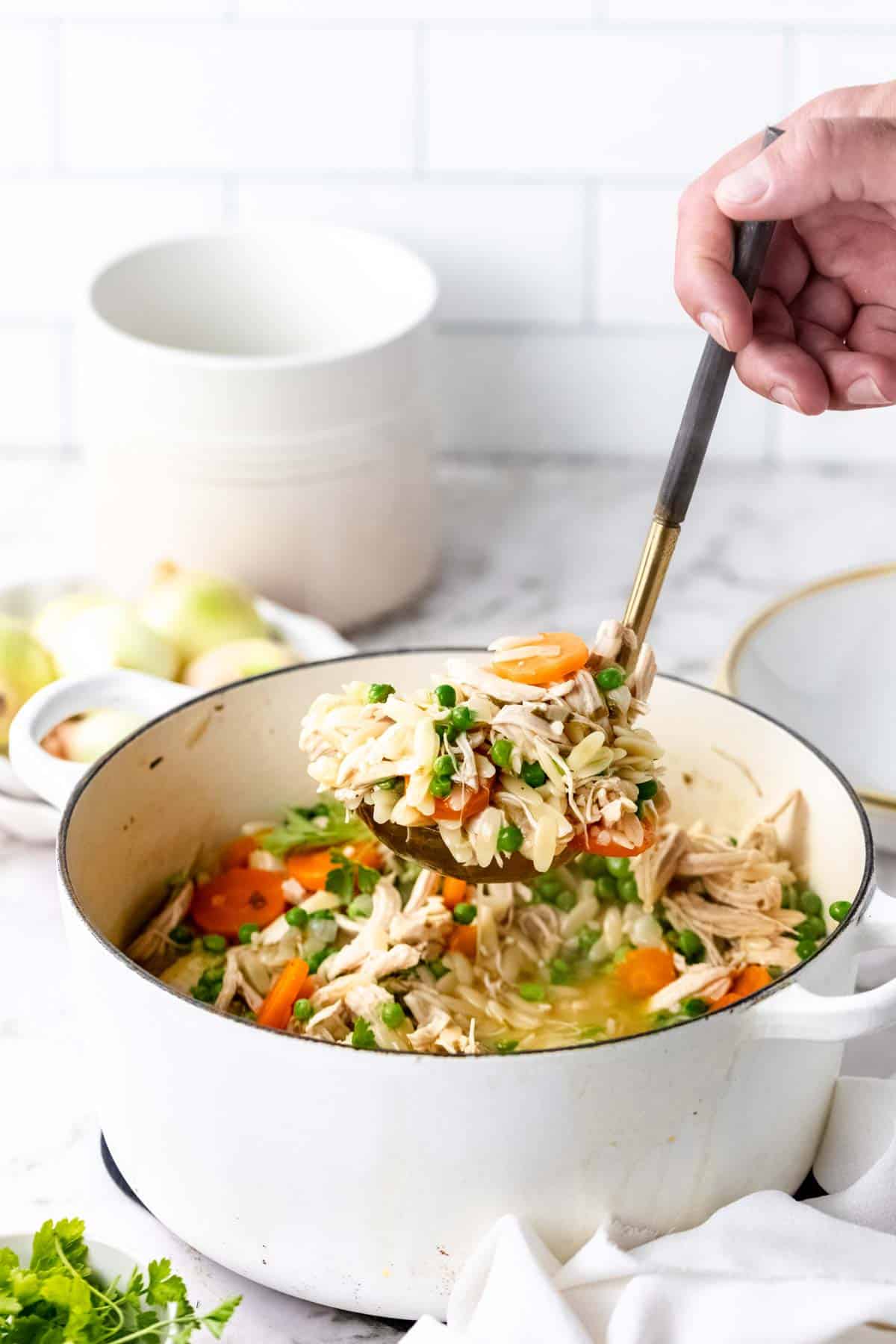 A hand holds a ladle filled with chicken orzo soup above a white pot on a marble coutertop.