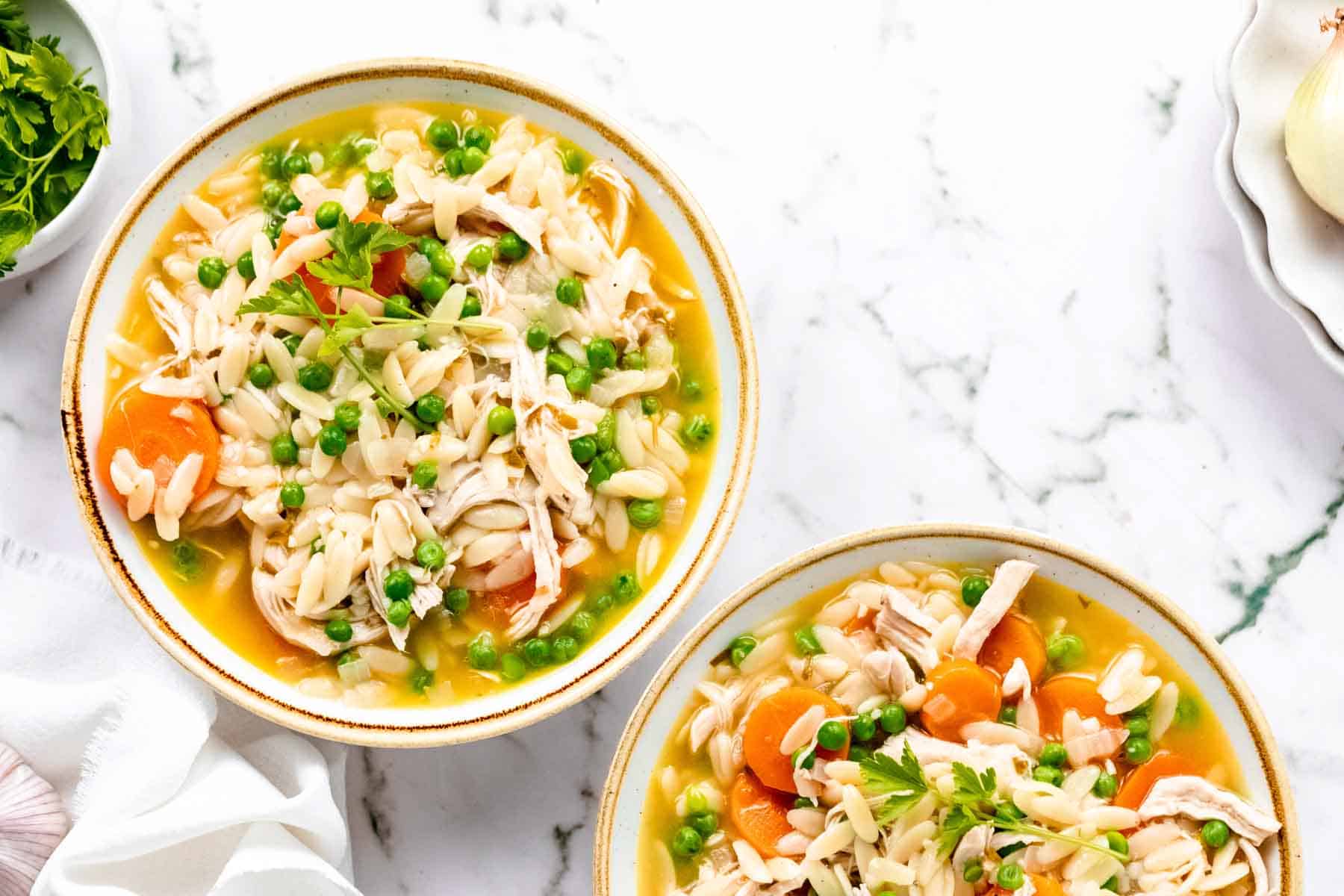 Overhead view of two bowls of gluten-free chicken orzo soup on a white marble countertop.