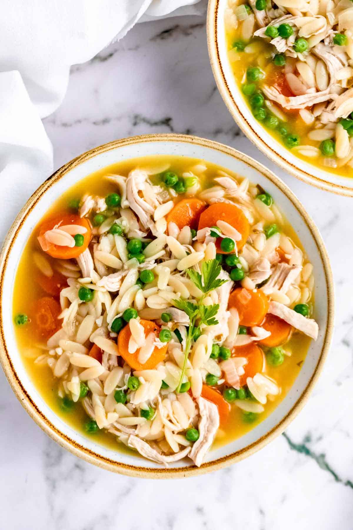 Overhead view of two bowls of gluten-free chicken orzo soup on a white marble countertop.