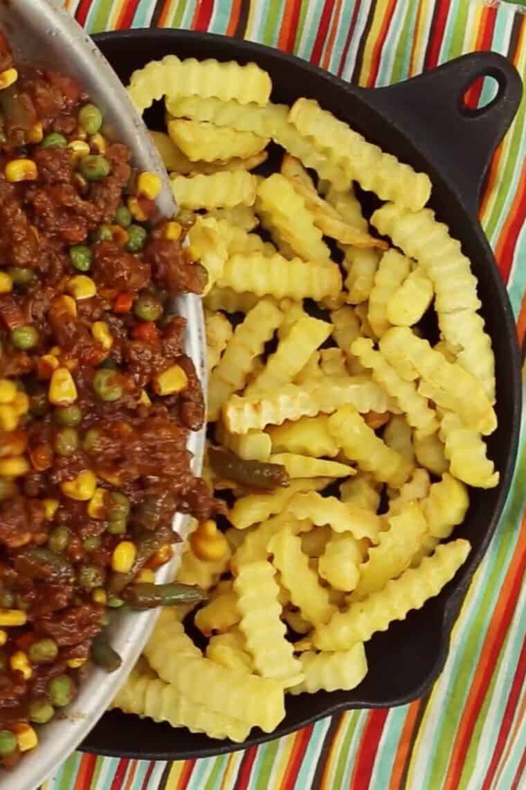 The shepherd's pie beef mixture being poured over baked crinkle cut fries in a cast iron skillet.