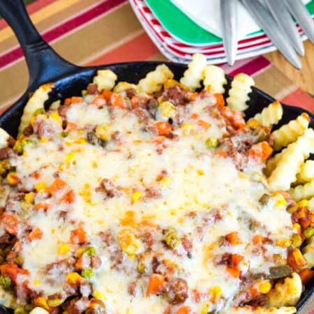 Overhead cast iron skillet with baked french fries topped with a shepherd's pie mixture of ground beef and mixed vegetables and melted cheddar cheese.