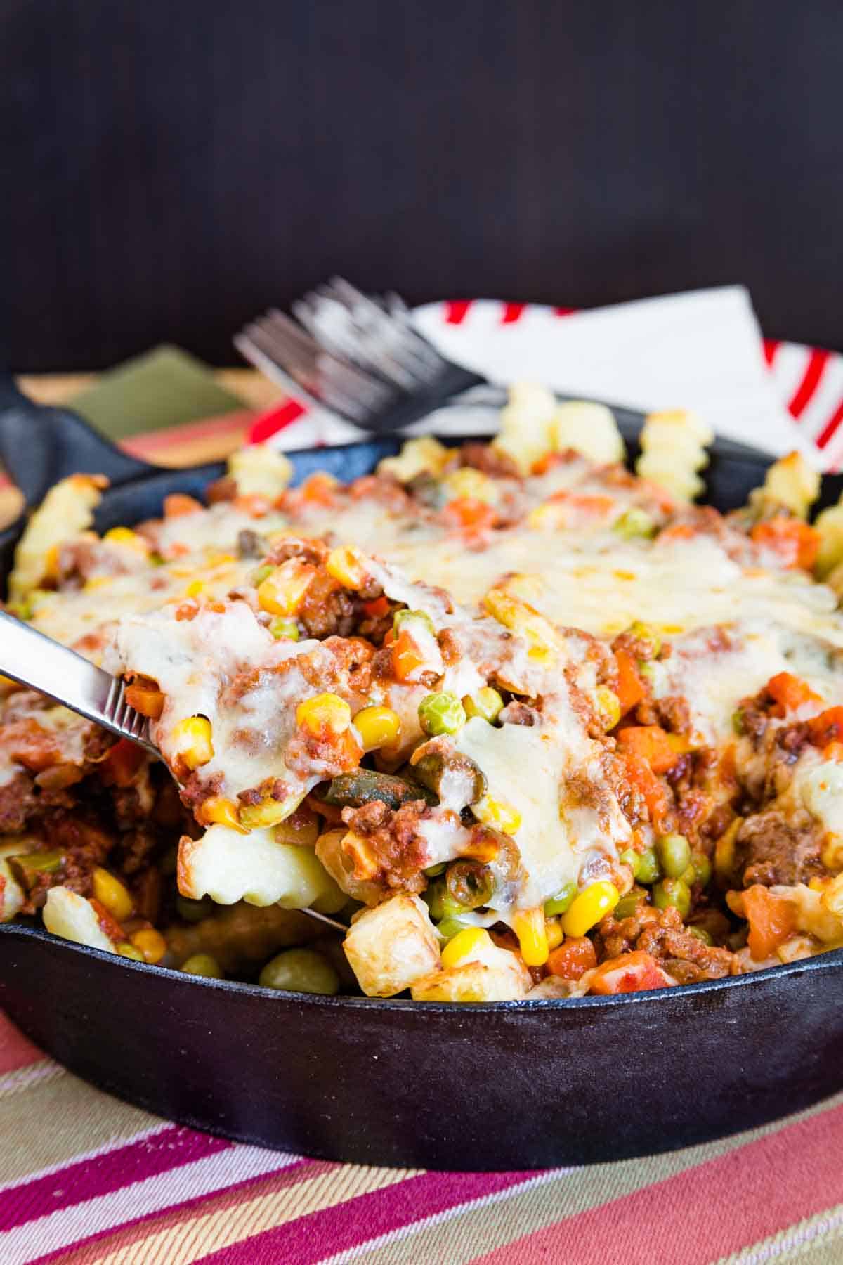 A spoon scooping up loaded french fries with ground beef, mixed vegetables, and cheese out of a cast iron skillet.