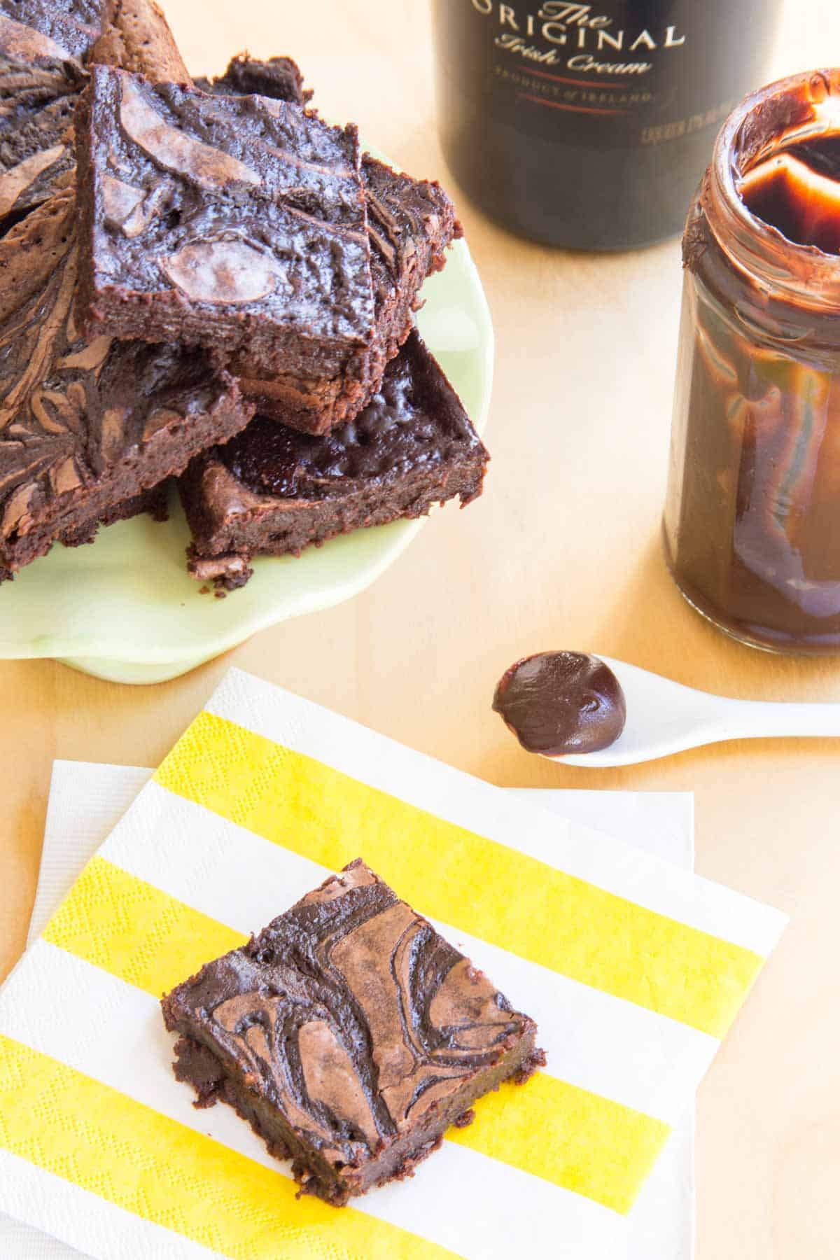 Overhead of a cake stand of Irish Cream Brownies with one Bailey's Brownie on a striped napkins, a jar of fudge sauce, and a bottle of Bailey's