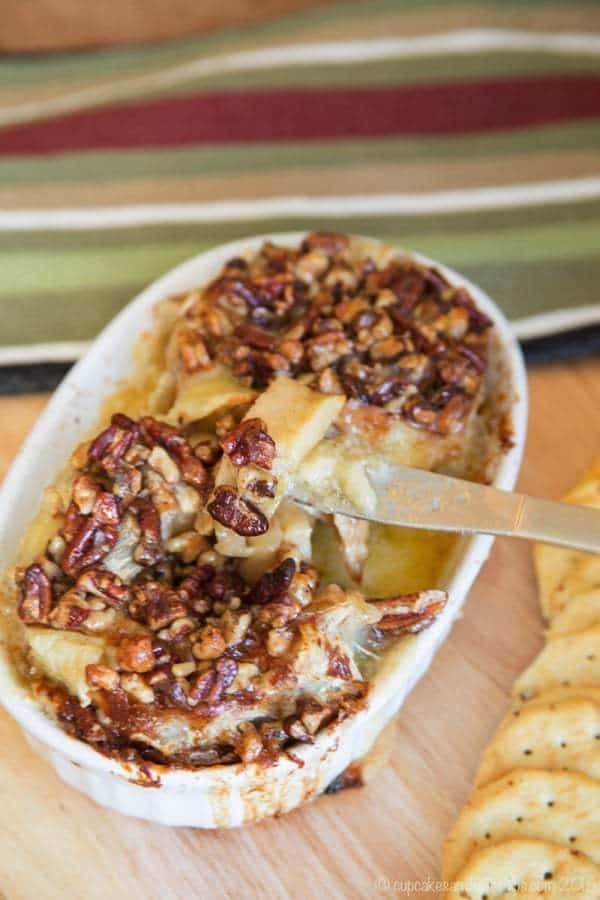 A cheese knife cuts into Honey Nut Baked Brie Bloomin' Apples served in a baking dish next to crackers