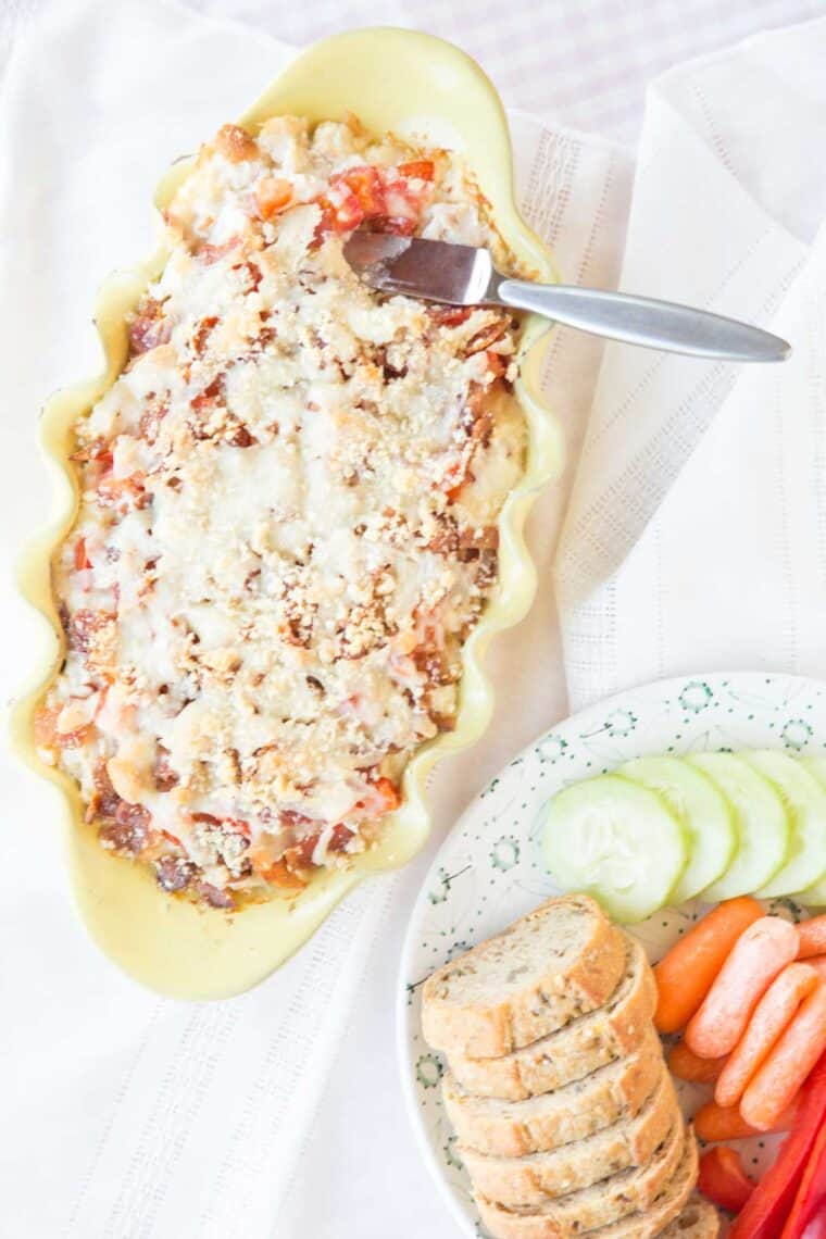 Top of a hot dip in a baking dish covered with cheese next to a plate of carrots, cucumbers, and bread slices