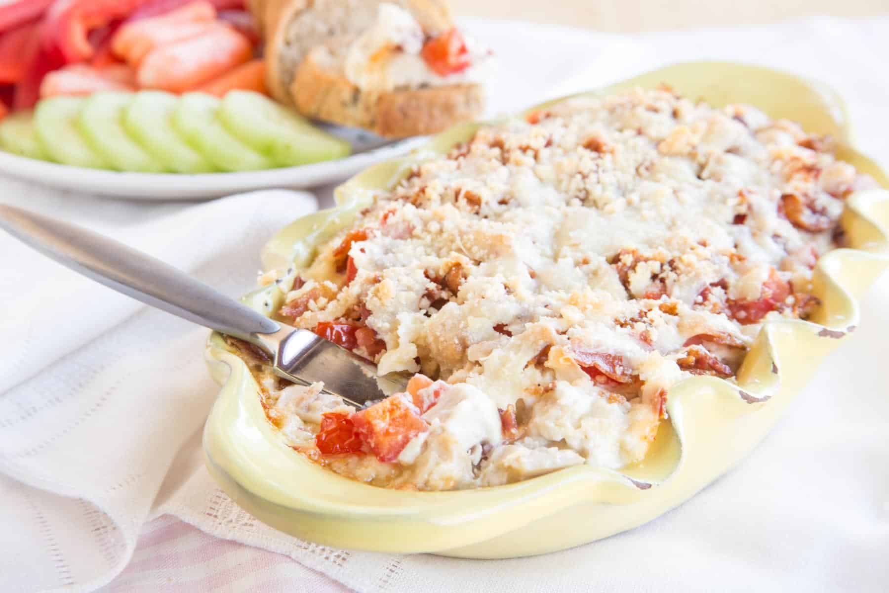 Hot Turkey, Bacon, and Cheese Dip served in a yellow fluted baking dish with a knife for spreading