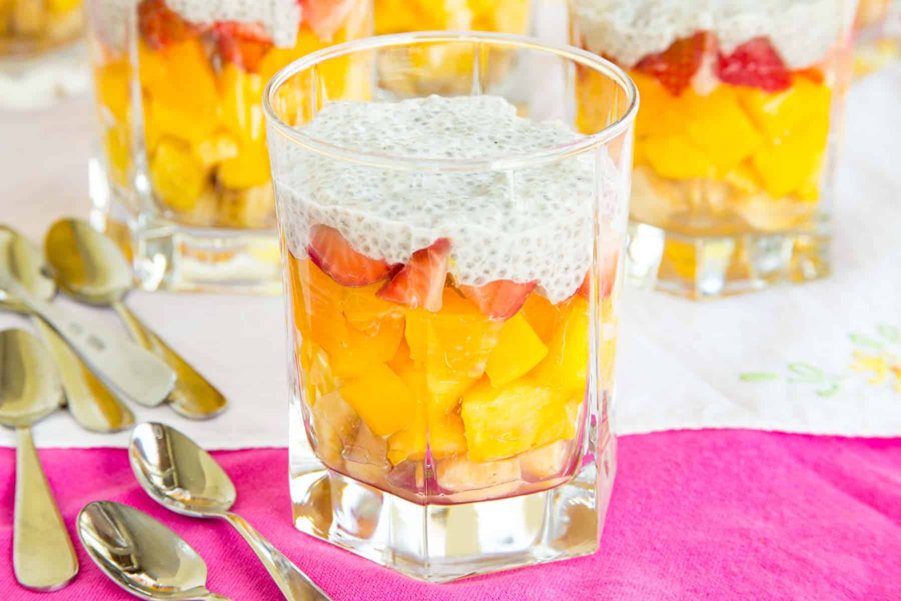 Tropical fruit layered in glasses with a coconut chia seed topping in glasses on a table with several mini spoons.