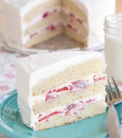 A slice of gluten free strawberries and cream cake on a turquoise plate with a fork and the whole cake in the background.