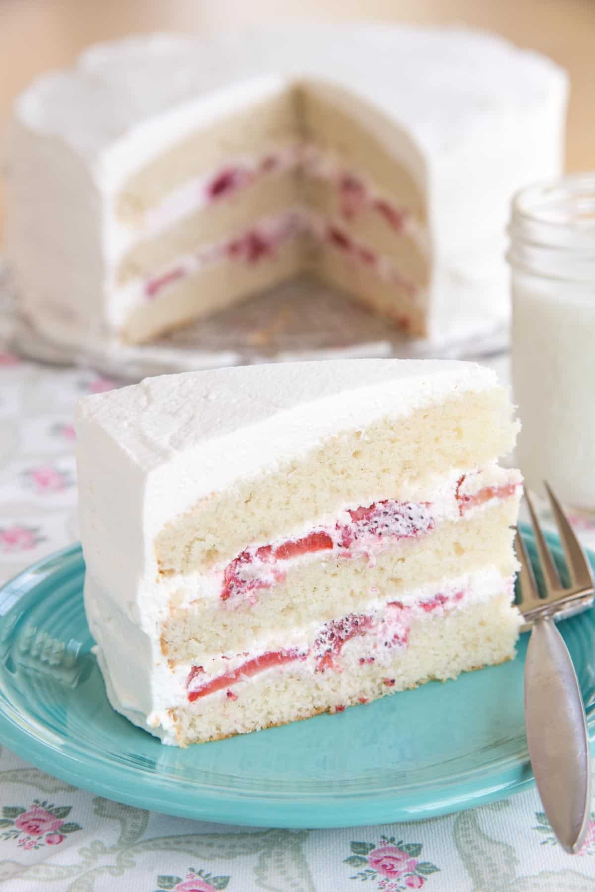 A slice of gluten free strawberries and cream cake on a turquoise plate with a fork and the whole cake in the background.