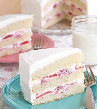 A slice of gluten free strawberries and cream cake on a turquoise plate with a fork with another piece on a pink plate and the whole cake in the background.