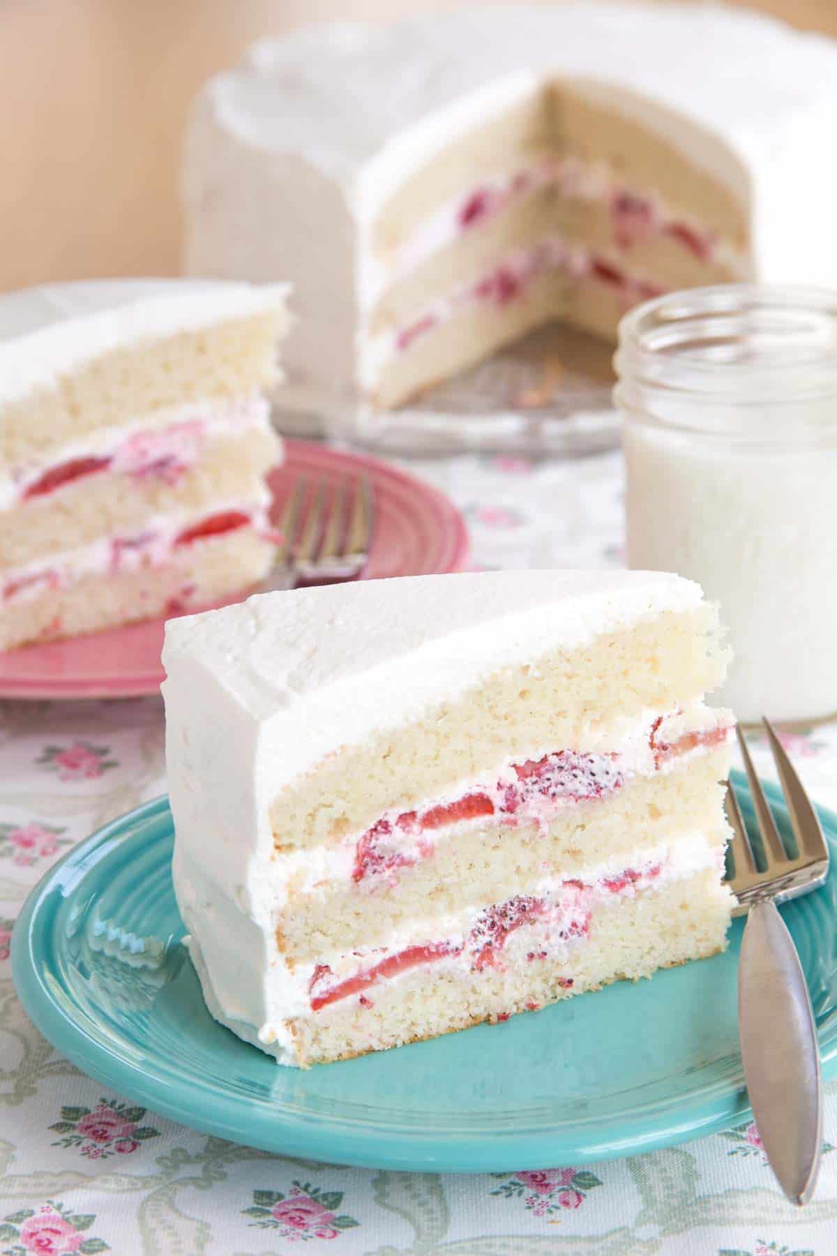 A slice of gluten free strawberries and cream cake on a turquoise plate with a fork with another piece on a pink plate and the whole cake in the background.