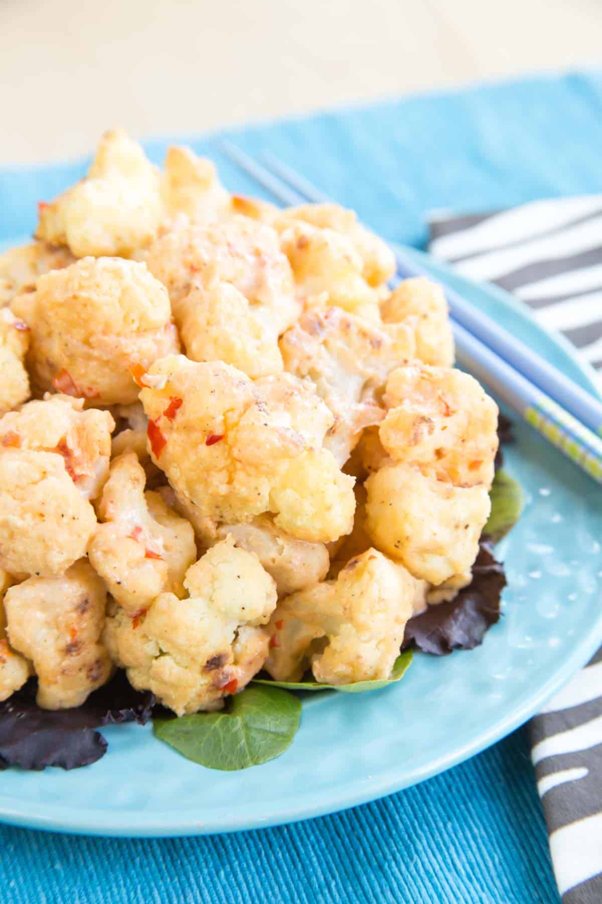 A plate of cauliflower bites covered in bang bang sauce on top of a blue placemat with a pair of chopsticks.