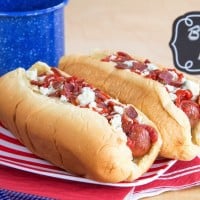 Two hot dogs in buns topped with buffalo ketchup, bacon, and crumbled blue cheese