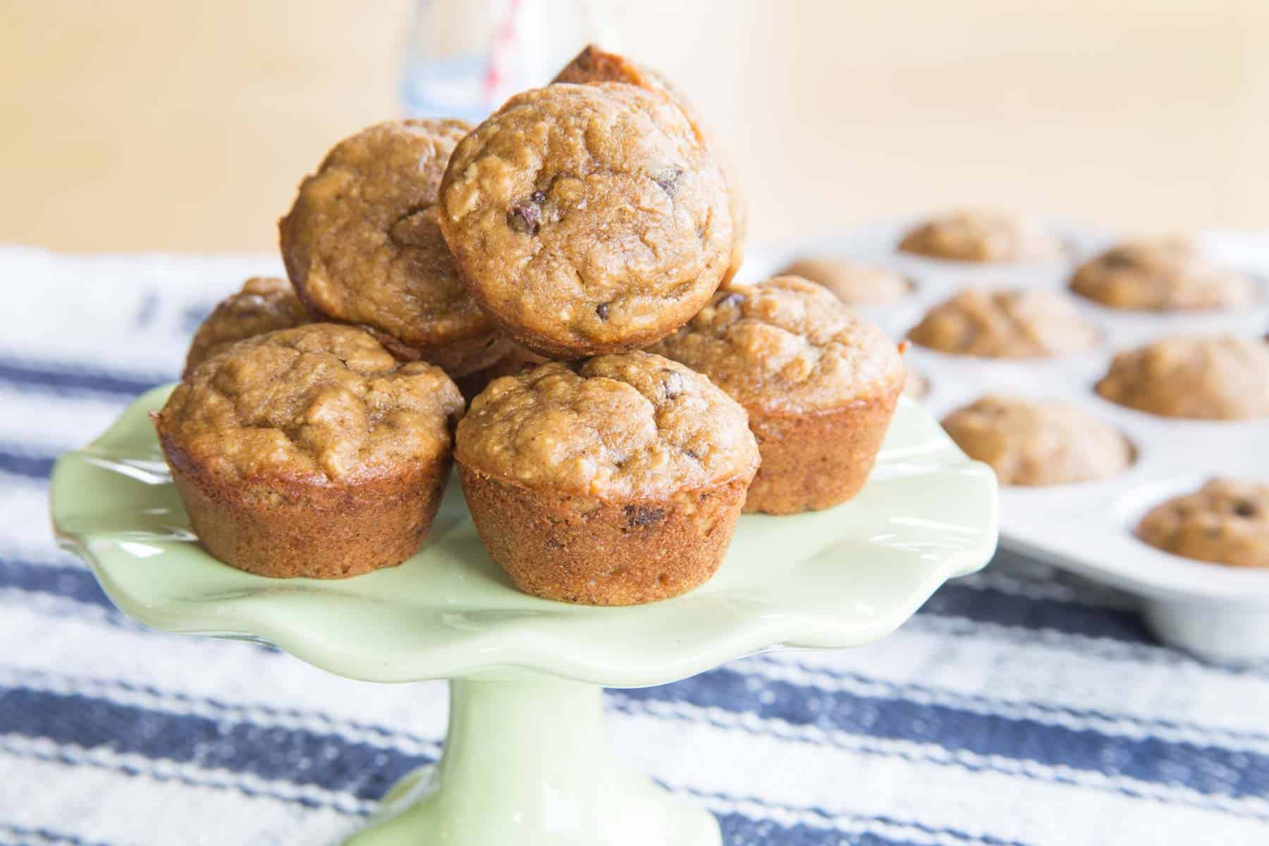 Gluten Free Peanut Butter Banana Muffins piled on a small cake stand with more mini muffins in a muffin pan in the background.
