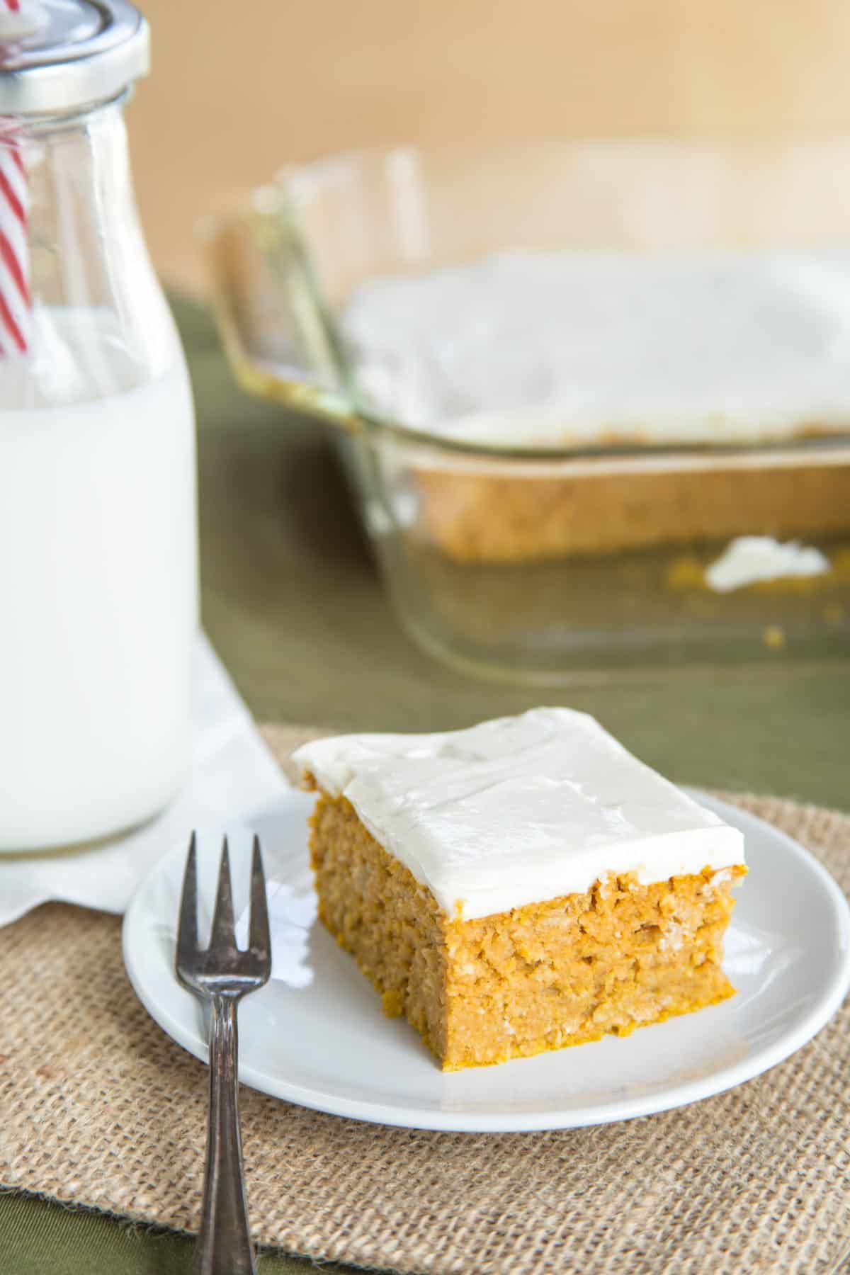 Pumpkin snack cake served on a plate, topped with Greek yogurt cream cheese frosting next to a glas bottle of milk and the rest of the cake in a pan