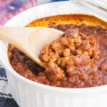 A wooden spoon picking up a scoop of baked beans out of a white casserole dish with text overlay that says "Semi-Homemade Baked Beans".