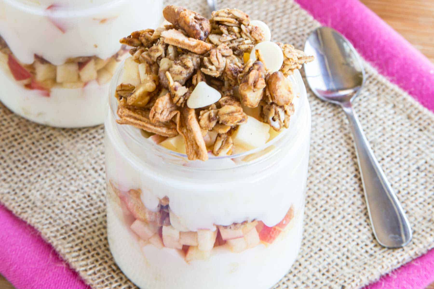 An apple parfait with layers of yogurt, apples, and granola on a piece of burlap and a pink cloth napkin.