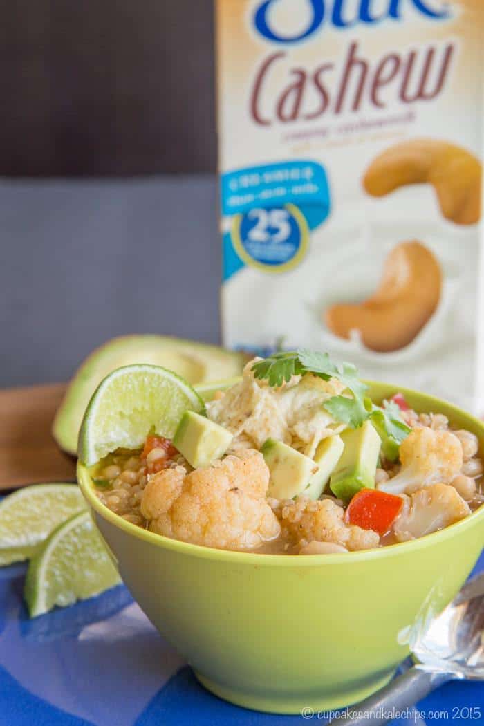 A bowl of quinoa chili next to a spoon and lime wedges, with a carton of Silk cashew milk in the background.