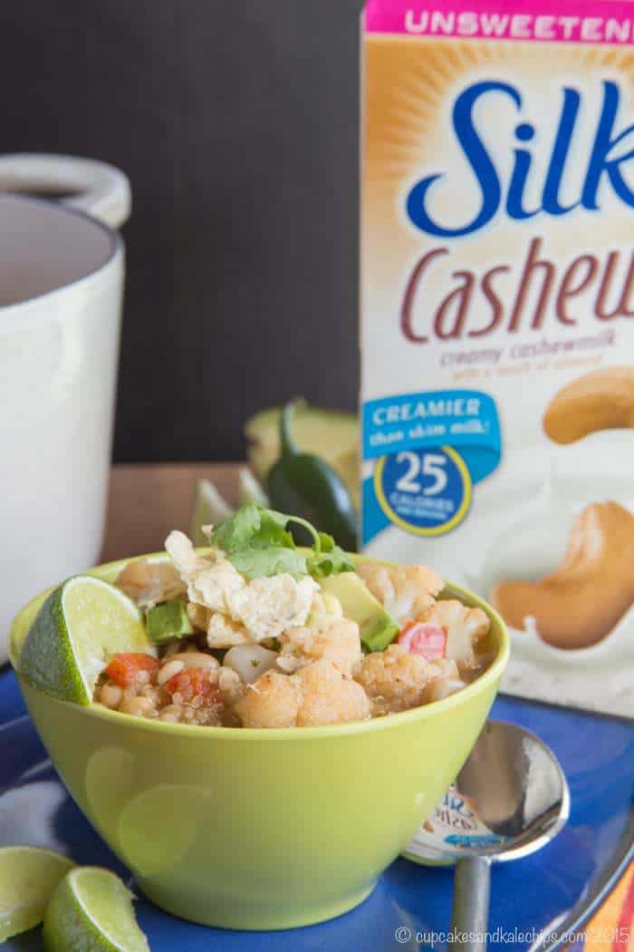 A bowl of quinoa chili next to a spoon and lime wedges, with a large pot and a carton of Silk cashew milk in the background.