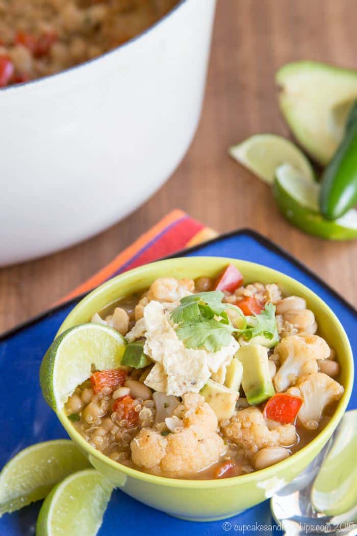 A bowl of quinoa chili next to a spoon and lime wedges, with a large pot in the background.