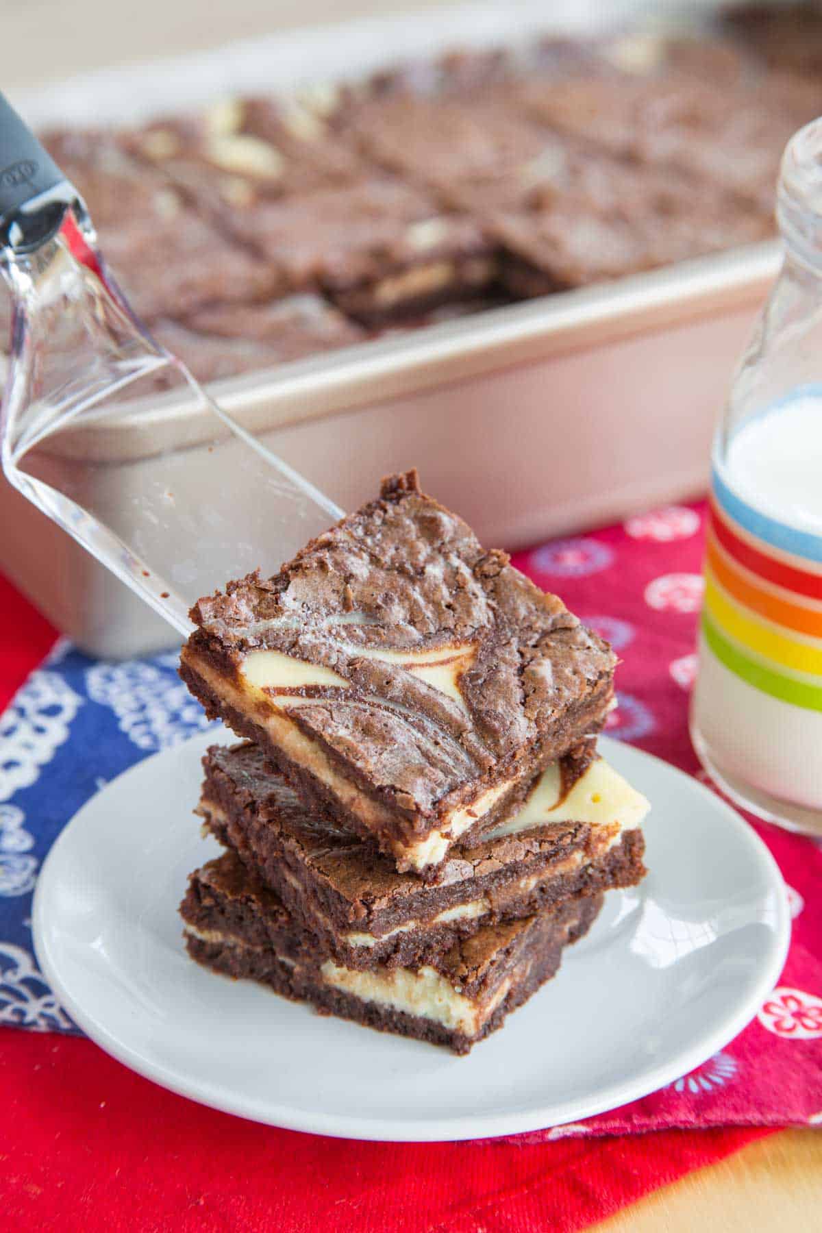 A spatula adding a brownie to the top of a stack on a plate with the whole pan in the background.