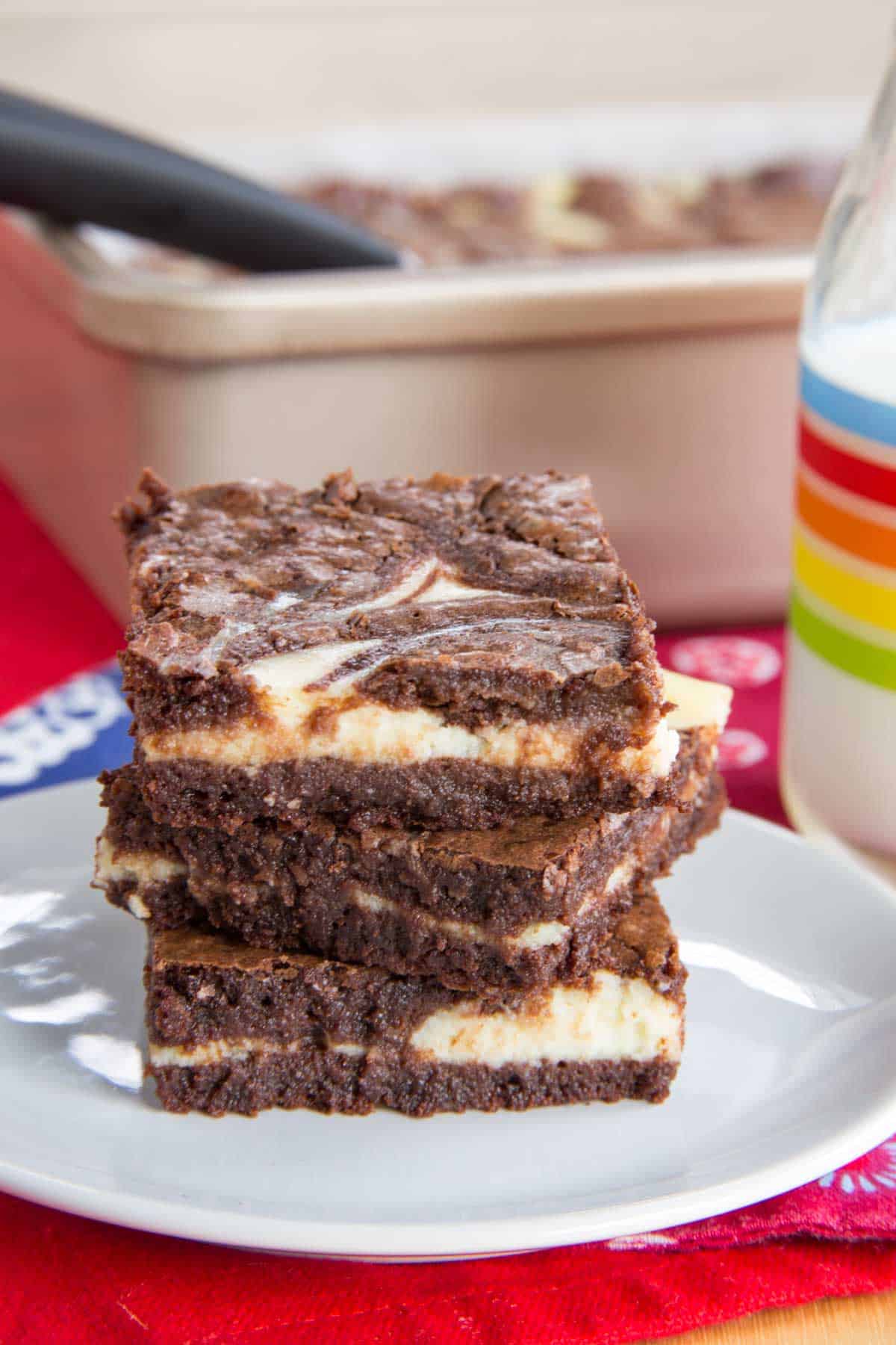 Three cheesecake brownies on top of each other on a small white plate on a red napkin.