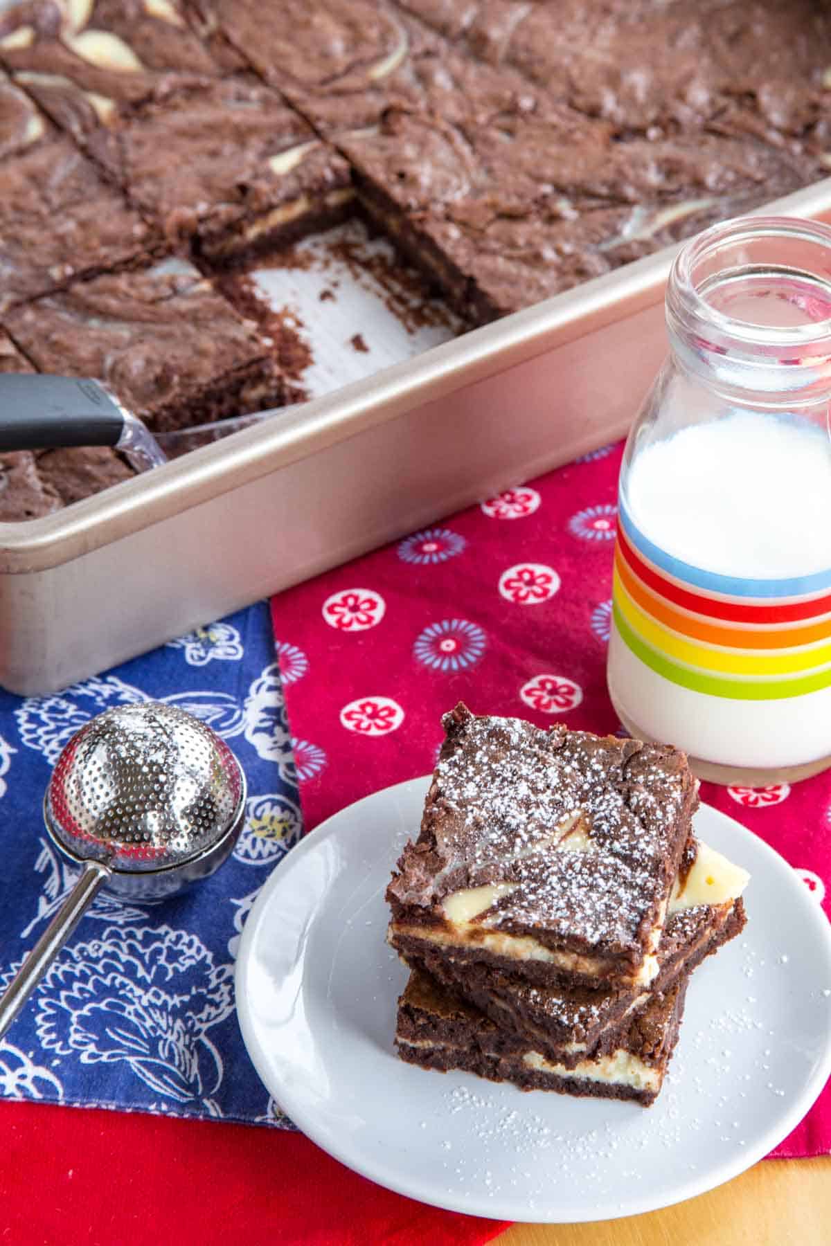 Powdered-sugar dusted brownies on a plate with the rest of the pan of brownies behind it.