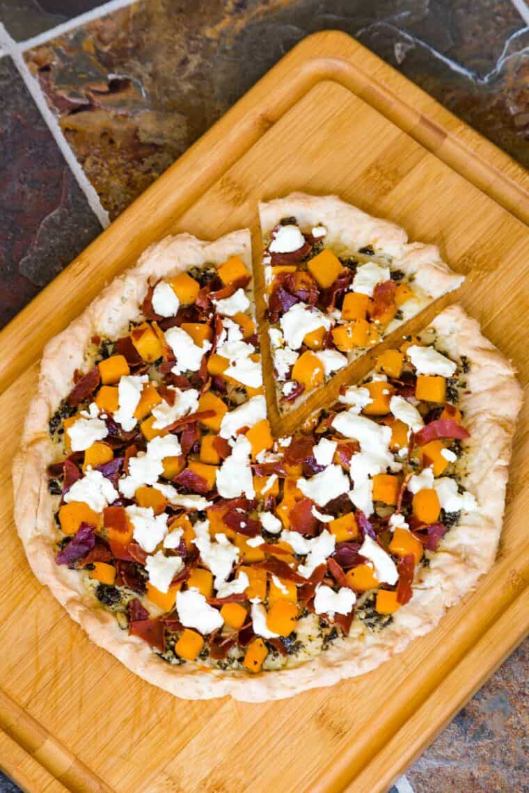 Looking down at a pizza topped with pesto, roasted butternut squash, goat cheese, and prosciutto on a cutting board.
