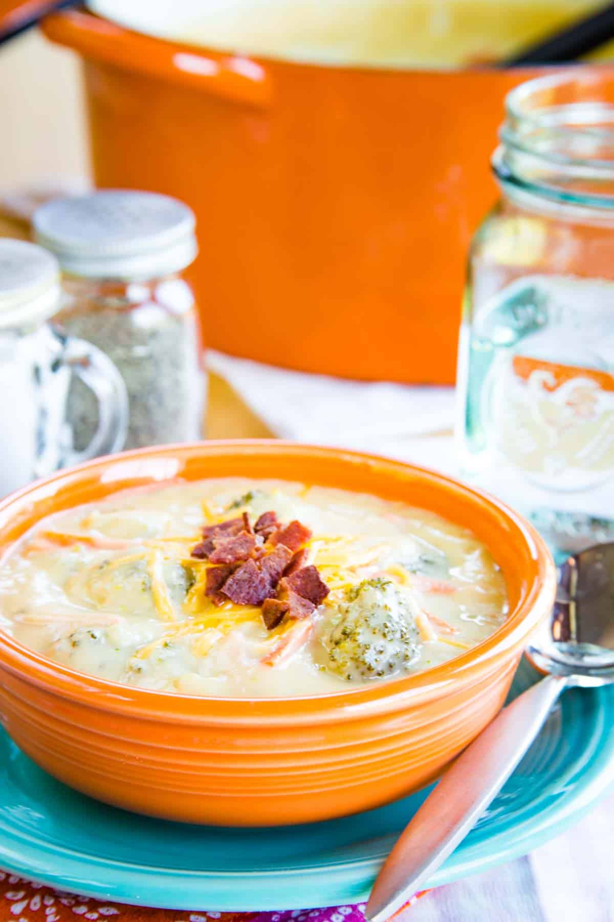 An orange bowl on top of a turquoise plate filled with broccoli cheddar soup with a pot and a salt and pepper shaker in the background.