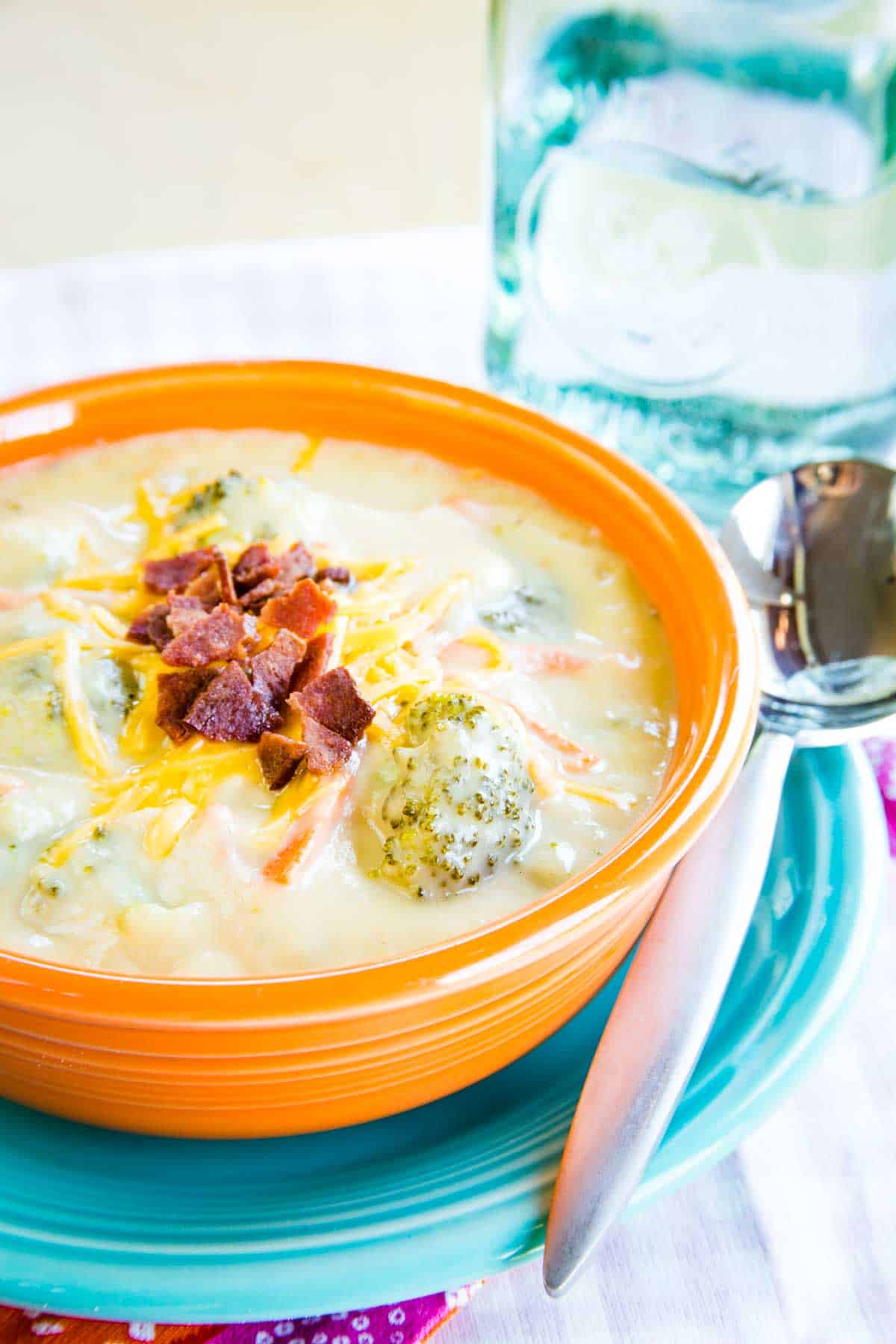 A bowl of broccoli cheese soup and a soup spoon on top of a small turquoise plate with a glass of water.