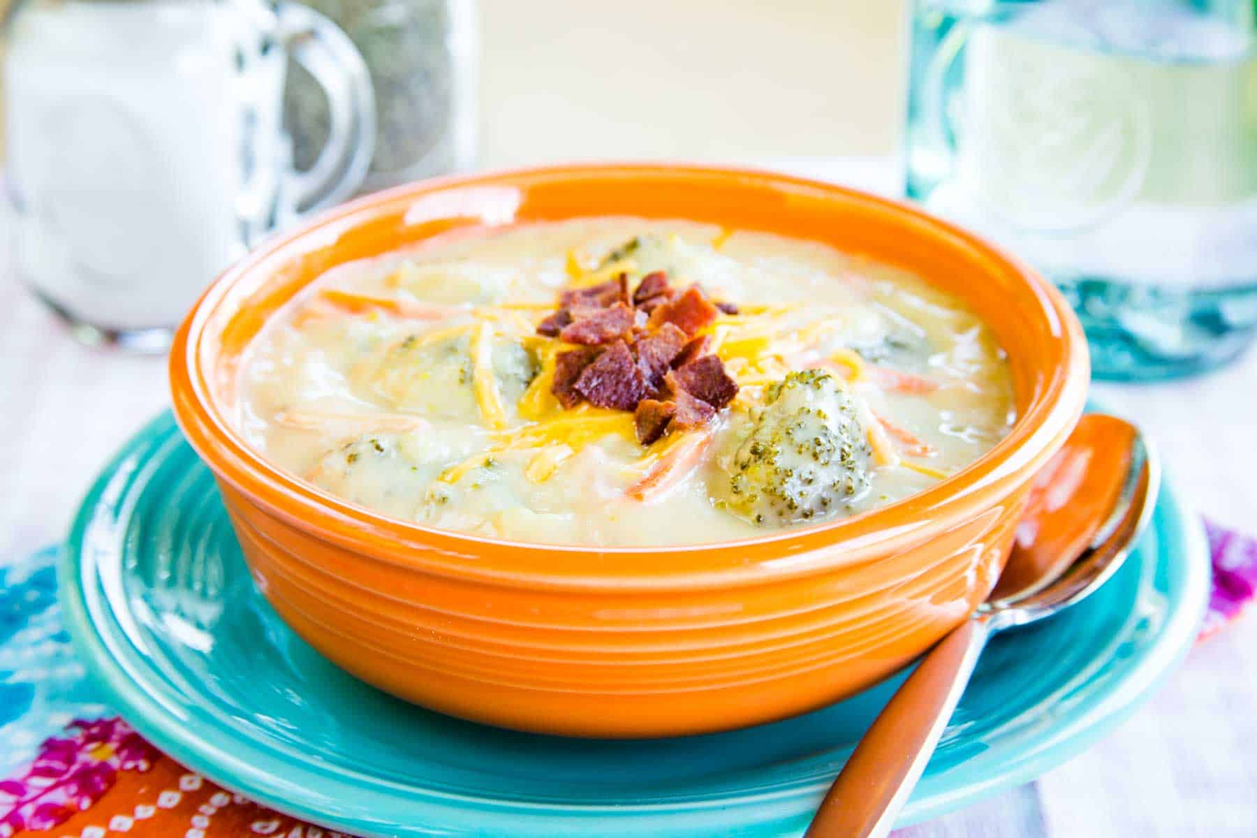 An orange bowl of keto Cauliflower Broccoli Cheese Soup set on top of a turquoise plate topped with crumbled bacon.