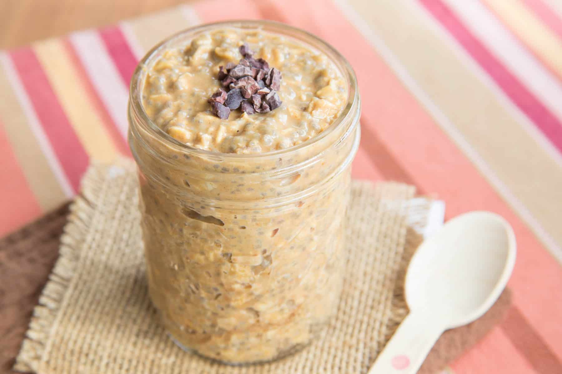 A glass jar of Pumpkin Spice Latte Overnight Oats with a spoon next to it on top of a piece of burlap and a striped placemat.